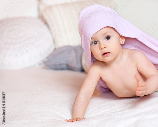 Fototapeta A cute baby after bathing in a bright room on a white bed stands on his hands. Pink baby towel. Baby's first steps. Textiles and bedding for children. Family morning at home.