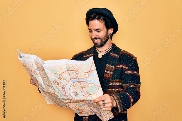 Obraz Young handsome hispanic bohemian hipster man with bear looking a tourist city map with a happy face standing and smiling with a confident smile showing teeth