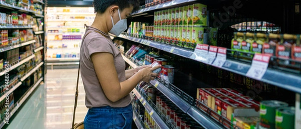 Fototapeta Asian woman wearing face mask selecting foods in supermarket to buy during coronavirus crisis or covid19 outbreak. Water supplies, grocery, prepare food, social distancing or new normal concepts