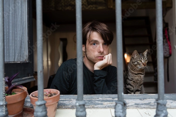 Obraz Young man looking through a barred window with his cat