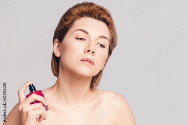 Fototapeta a beautiful model - a brown-haired woman sprays a perfume from a red bottle on her neck. Shot on a gray background in the studio. Closeup face of young beautiful woman with a healthy clean skin