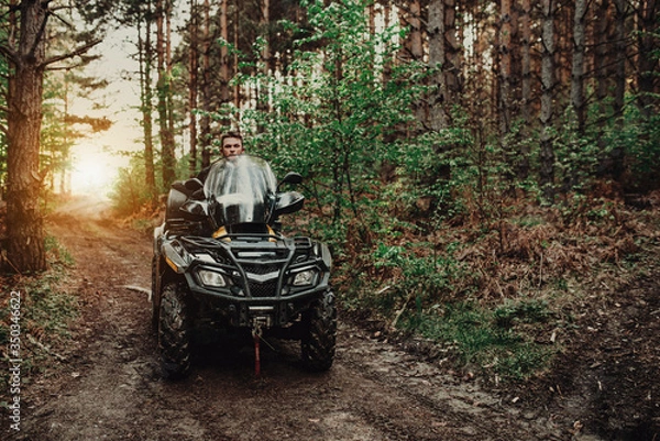 Fototapeta A young man in a white helmet rides through the woods on a Quad bike. Extreme hobby. A trip to ATV on the road from logs. Quad Biking through the forest.