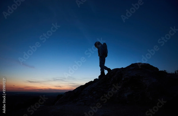 Fototapeta Beautiful view of space traveler on rocky mountain with majestic night sky on background. Silhouette of cosmonaut in space suit exploring new planet. Concept of space travel and twilight beauty.