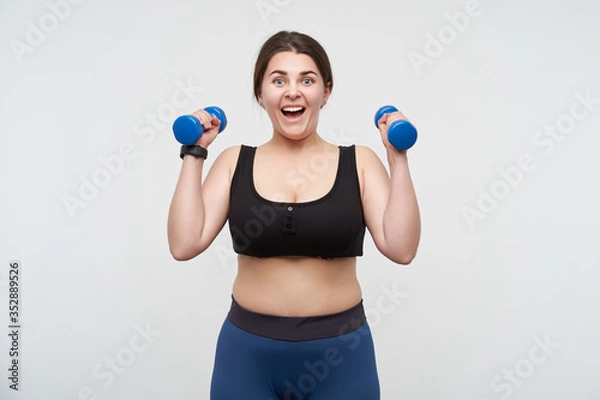 Obraz Agitated young cheerful oversized woman with casual hairstyle looking emotionally at camera while raising hands with blue dumbbells, posing over white background. Body positive concept