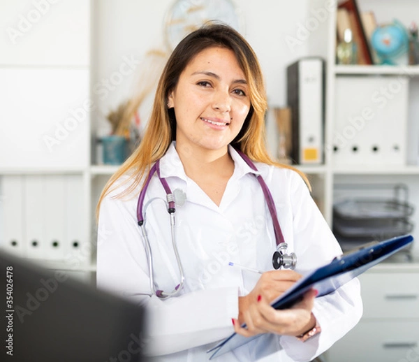 Fototapeta Female doctor in medical coat holding clipboard in clinic