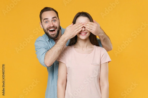 Fototapeta Excited young couple two friends guy girl in pastel blue casual clothes posing isolated on yellow background studio portrait. People lifestyle concept. Mock up copy space. Covering eyes with hands.