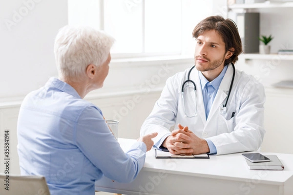 Fototapeta Attentive doctor listening to senior patient at his office