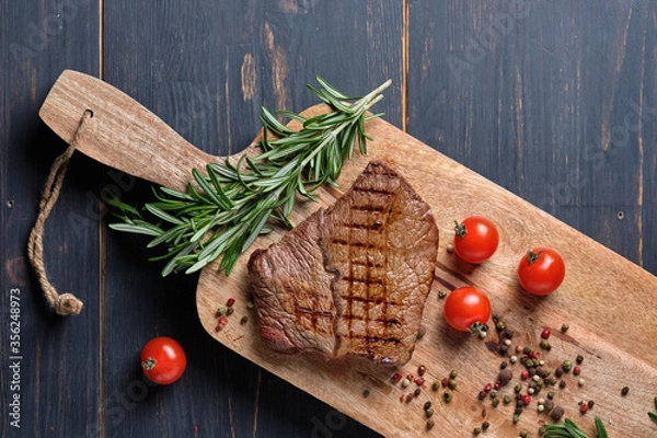 Fototapeta Freshly cooked steak on a wooden board with rosemary and cherry tomatoes. Close-up. Top view.