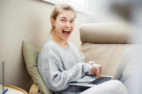 Fototapeta Cheerful young blonde looking at side holding laptop in her hands sitting in chair