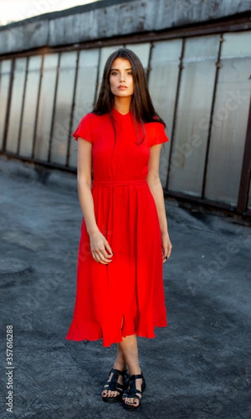 Fototapeta Attractive beautiful shy girl in a red dress with brown straight hair, looking at camera. Full length portrait.