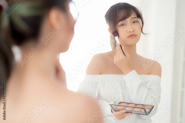 Fototapeta Beauty portrait young asian woman smiling with face looking mirror applying makeup with brush cheek in the bedroom, beautiful girl holding blusher, skin care and cosmetic table fashion concept.