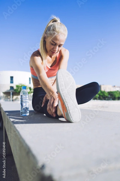 Fototapeta Portrait of a beautiful athletic female stretching legs muscles while sitting on concrete pter in summe summer day, young fit woman doing warm up exercises before begin morning run in the fresh air