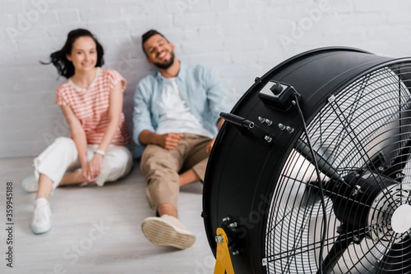 Fototapeta Selective focus of electric fan near smiling couple sitting on floor at home