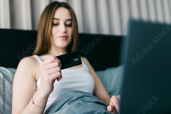 Fototapeta Beautiful young woman using her credit card on the web shopping lying on the bed