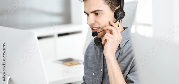 Fototapeta Casual dressed young man using headset and computer while talking with customers online. Call center, business concept