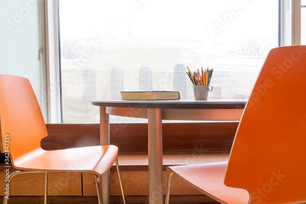 Fototapeta Desk with book and pencils at window. 2 orange chairs.