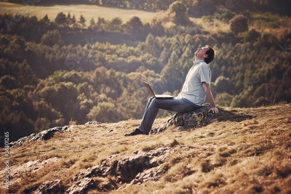 Fototapeta young man using laptop sitting on the grass on the hillside