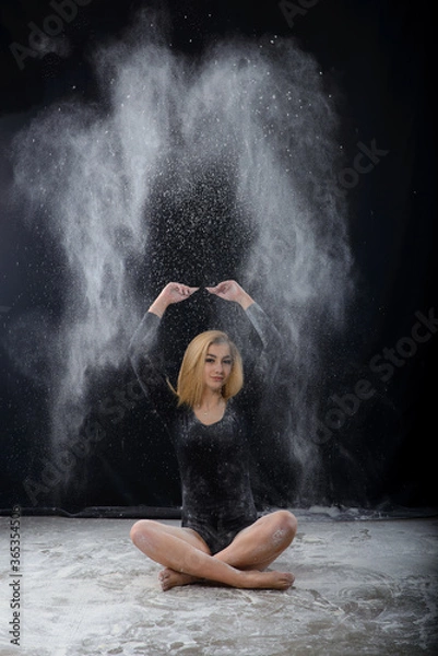 Fototapeta Beautiful plump blonde girl wearing a black gymnastic bodysuit covered with clouds of the flying white powder jumps dancing sitting on a dark. Artistic conceptual and advertising photo.