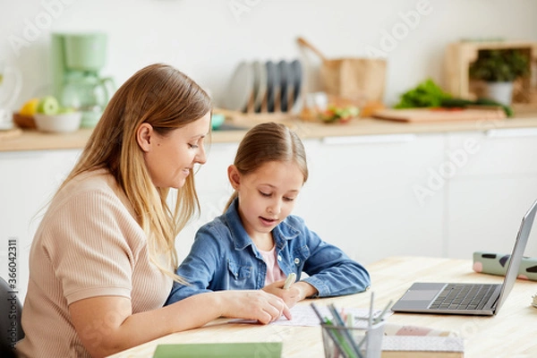 Fototapeta Warm-toned portrait of caring mother helping cute girl doing homework or studying at table in cozy home interior, copy space