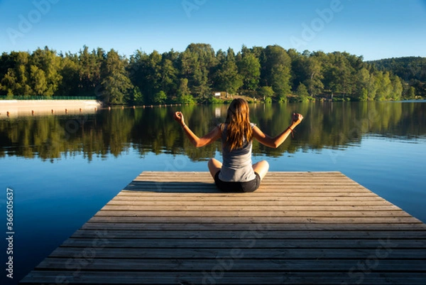 Fototapeta young woman on wooden pontoon or pier  practicing yoga & relaxing on holiday