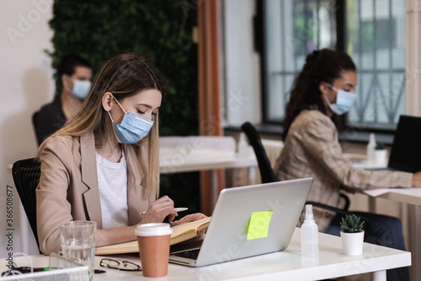 Fototapeta Portrait of a young female freelancer with mask working in an office while sitting at a table with colleagues in the background.