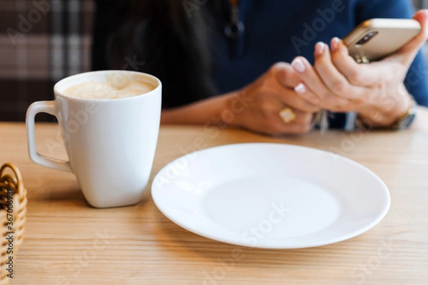 Obraz A white coffee Cup and an empty plate on a wooden table against the background girl's hands holding a smartphone while waiting for breakfast in the morning.