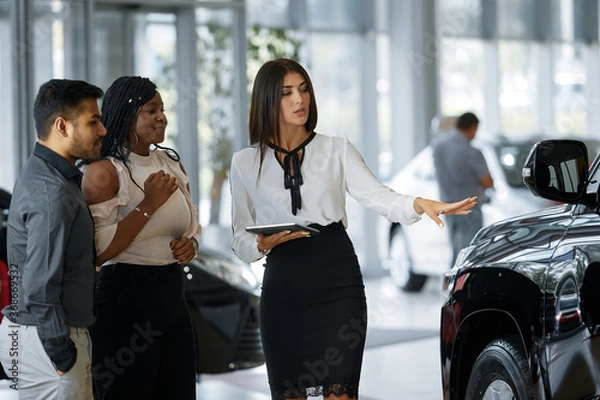 Obraz A beautiful couple smiles while looking at a new car in a car dealership with a seller. An agent demonstrates the new features of a luxury car to an African-American happy couple at a car dealership.