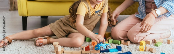 Obraz cropped view of babysitter near child playing with toy car near multicolored blocks on floor, horizontal image