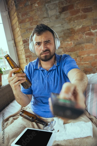 Fototapeta Bored using TV remote, drinks beer. Lazy man living in his bed surrounded with messy. No need to go out to be happy. Using gadgets, watching movie and series, looks emotional. Tablet with copyspace