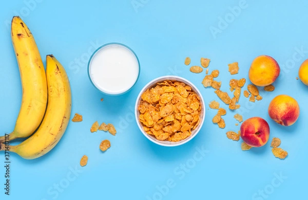 Fototapeta Breakfast cereals, bananas, apricots and a glass of milk on a bright blue background top view, flat lay. Fitness food concept. Breakfast with cornflakes, milk and fruit. Copy space.