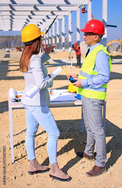 Fototapeta Female architect and construction engineer in hardhats talking about the project on construction site, behind them construction worker with measuring device, crane and construction workers, teamwork