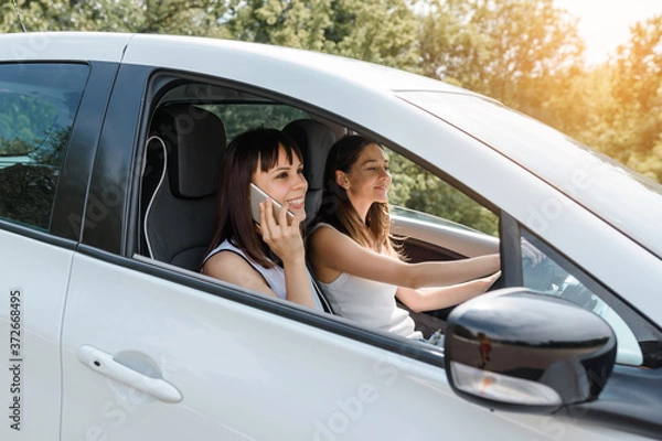 Fototapeta Two happy young women friends are driving a car and enjoy the company.