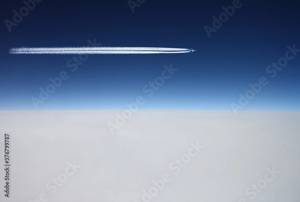 Fototapeta Airplane flys above white clouds in a deep blue sky and leaving jet trail