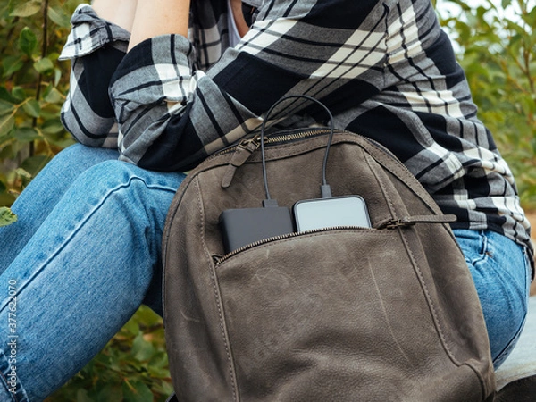 Fototapeta The phone is charged using a power Bank. Use of modern technologies in everyday life. The phone and powerBank are in the tourist's leather backpack. Convenient modern gadgets for a comfortable life