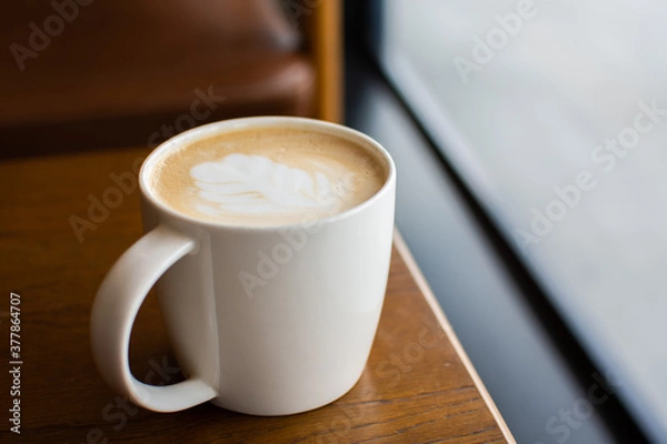 Fototapeta White coffee cup with foam latte art on a brown wooden table in the coffee shop