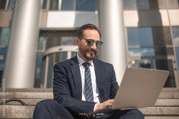 Fototapeta Successful businessman works on laptop sitting on steps of business center. Smiling man wearing business suit and sunglasses. High quality photo.