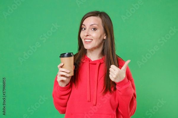 Obraz Smiling beautiful cheerful young brunette woman 20s wearing red casual streetwear hoodie posing showing thumb up hold paper cup of coffee or tea isolated on green color background studio portrait.