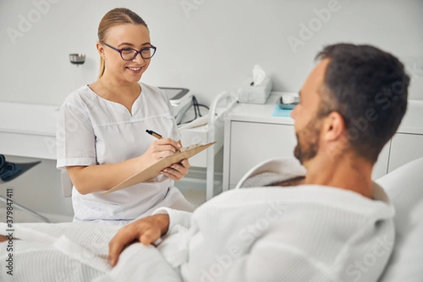 Fototapeta Cheerful beautician talking with male patient and taking notes