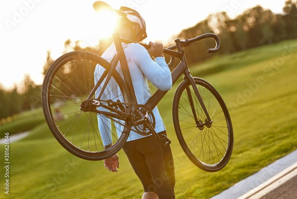 Obraz Strong athletic man in sportswear and protective helmet carrying his bicycle after cycling training in park, enjoying amazing sunset