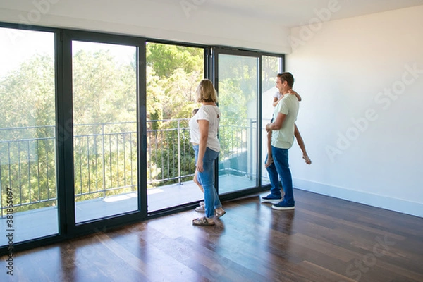 Fototapeta Happy family with kids looking at balcony and smiling. Back view of blonde mother. Daughter sitting on father hands. People watching new house or flat. Mortgage, relocation and moving day concept