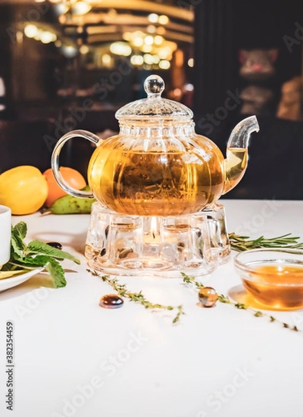 Fototapeta Fragrant fresh herbal tea and melissa in a glass teapot warming on a candle on a white table. Traditional herbal drink. Tea with honey in a restauran. Kettle warmer