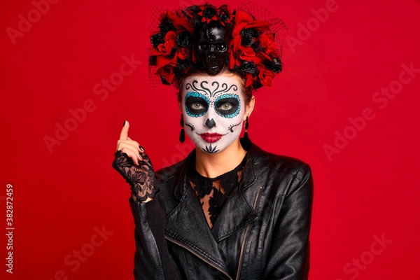 Fototapeta Scary young creepy lady calavera. wears artistic make-up for the feast of all the dead.Wears black leather jacket and lace gloves, dressed as skeleton isolated in red.