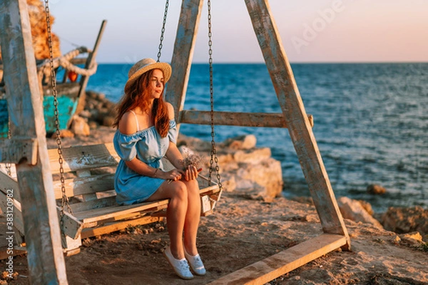 Fototapeta Romantic evening at sunset by the sea, a young woman in a dress with a bouquet of flowers is sitting on a wooden swing holding her hat