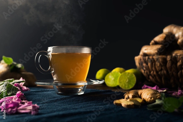 Fototapeta Immune boosting ginger and lemon tea with steam coming out of the cup and a basket full of ginger roots, lime, peppermint leaves and pink flowers. Black background with copy space.