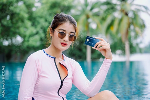 Fototapeta Asian woman traveler in swim suit hold a credit card at swimming pool outdoors.