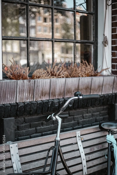 Fototapeta black and silver bicycle with a black bell stands near a large black window in which reflections of old Amsterdam houses are visible in blur and dry plants growing on the windowsill