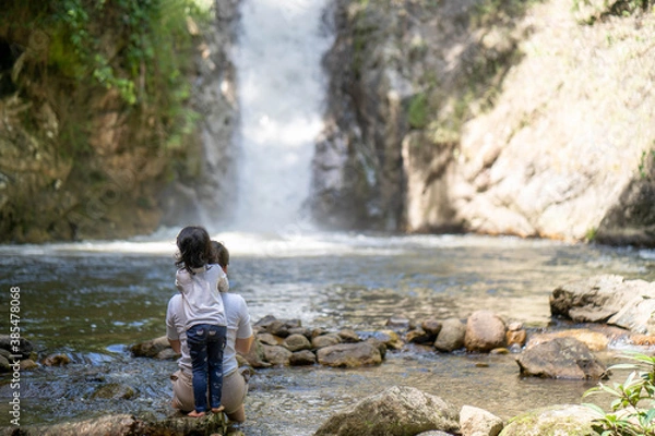 Fototapeta Mother and daughter sitting at the rocks against waterfall in woods forest.