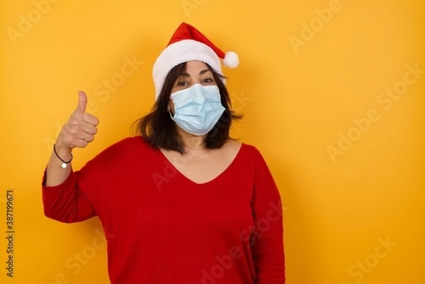 Fototapeta Beautiful middle aged woman wearing Christmas hat and medical mask standing against yellow background, pointing up with thumb. Protection against infectious disease.