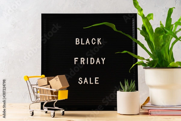 Fototapeta Text Black friday on black letter board, plants, shopping cart with boxes on wooden background. Concept season sales time. Template layout for your design.