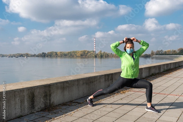 Fototapeta Young handsome healthy sporty fitness woman or girl stretching muscles before outdoor jogging and running training workout wearing protective medical mask on her face against coronavirus or COVID-19